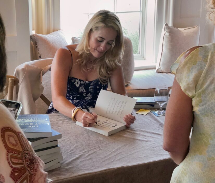 A woman sits at a table signing a book with a stack of books beside her. She is indoors near a window with people standing nearby.