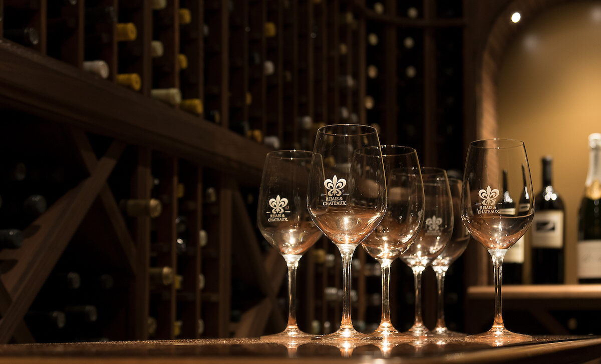 Five wine glasses sit on a wooden counter in a dimly lit wine cellar, with shelves of wine bottles in the background.