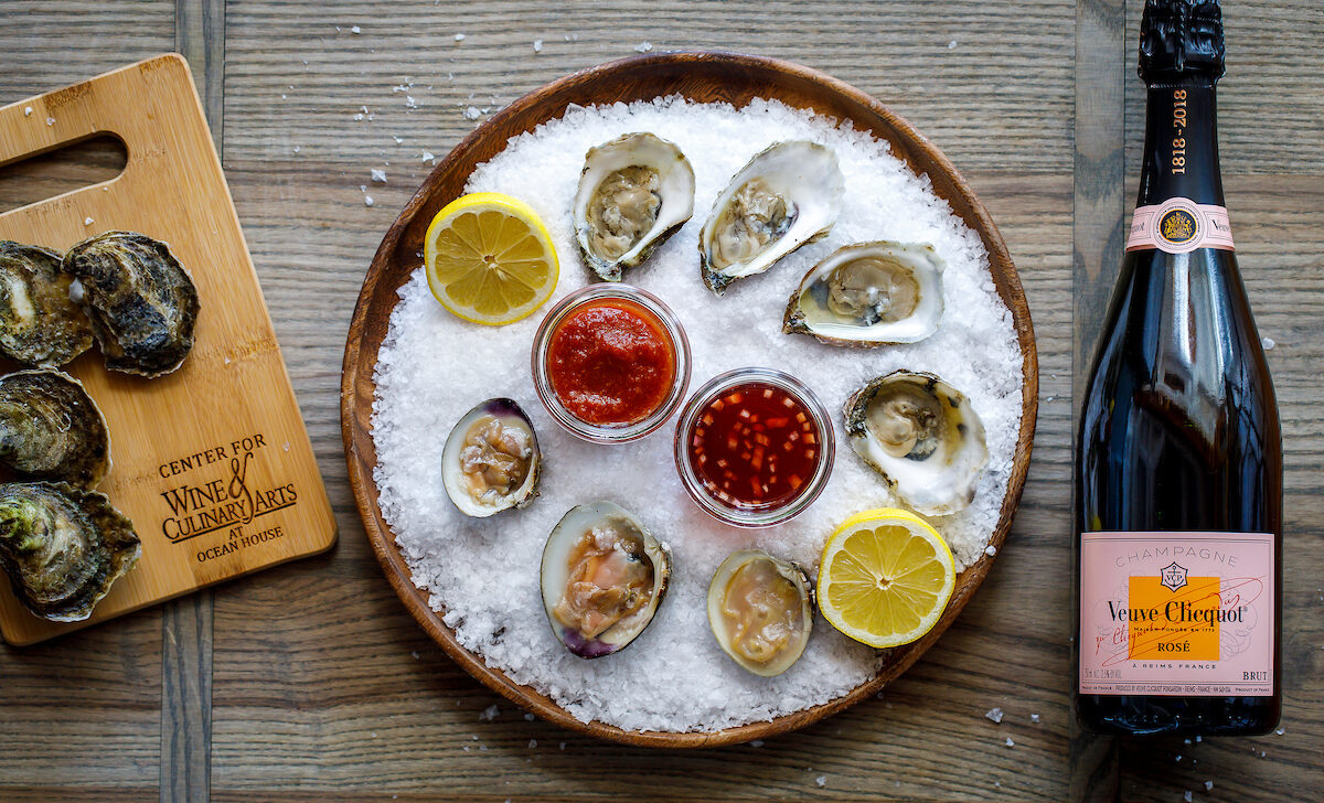 A platter of oysters on ice with lemon slices and dipping sauces sits beside a bottle of Veuve Clicquot Rosé champagne.