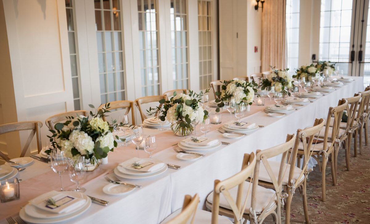 Elegant dining table set for a formal event with white plates, wine glasses, and floral centerpieces. The chairs are wooden, and the room is warmly lit with large windows in the background.