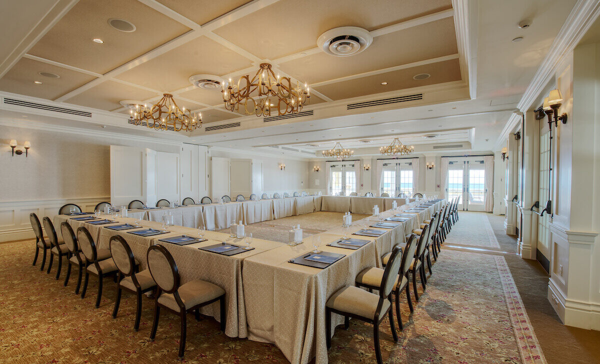 A large conference room with U-shaped table setup, chairs around, note pads, bottled water, carpeted floor, chandeliers, and windows overlooking a view.