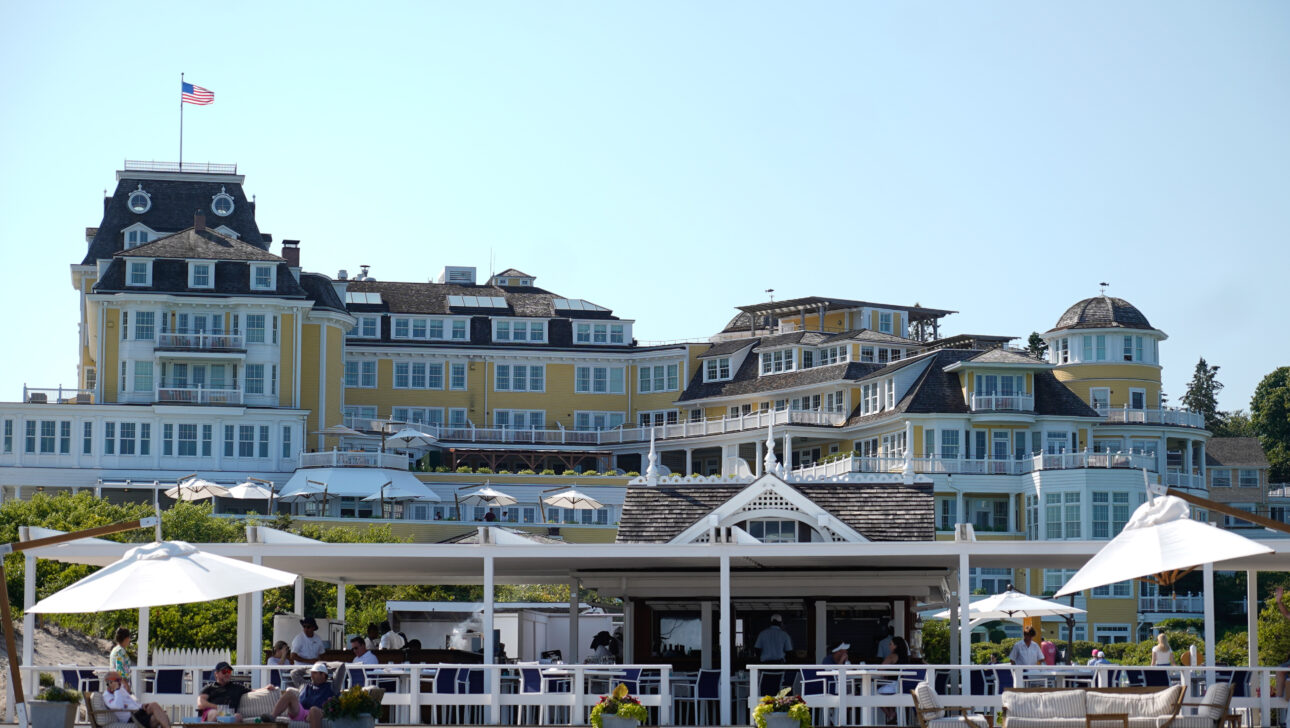 An ornate hotel with yellow and white siding is seen with a large structure in front, featuring outdoor seating and umbrellas. An American flag flies atop the hotel.