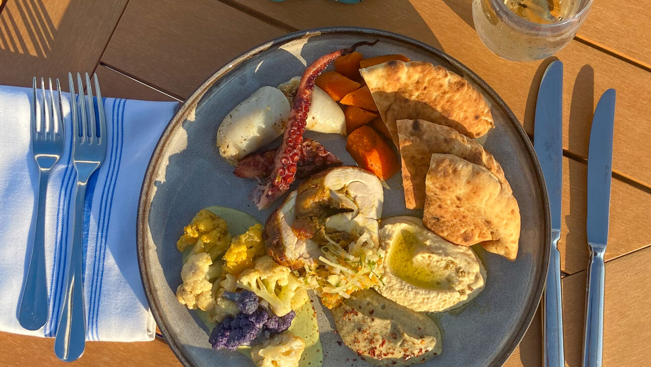 A dinner plate with grilled vegetables, pita bread, and chicken. Two forks and knives are on a napkin beside the plate. Glass of water and salt shakers are on the wooden table.