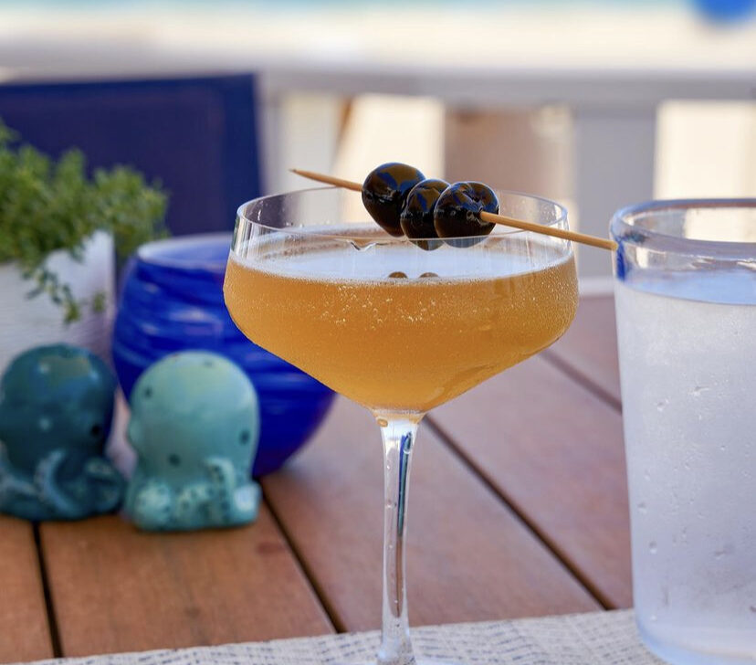 A cocktail garnished with two olives on a stick sits on a table, with a glass of water nearby and the ocean in the background.