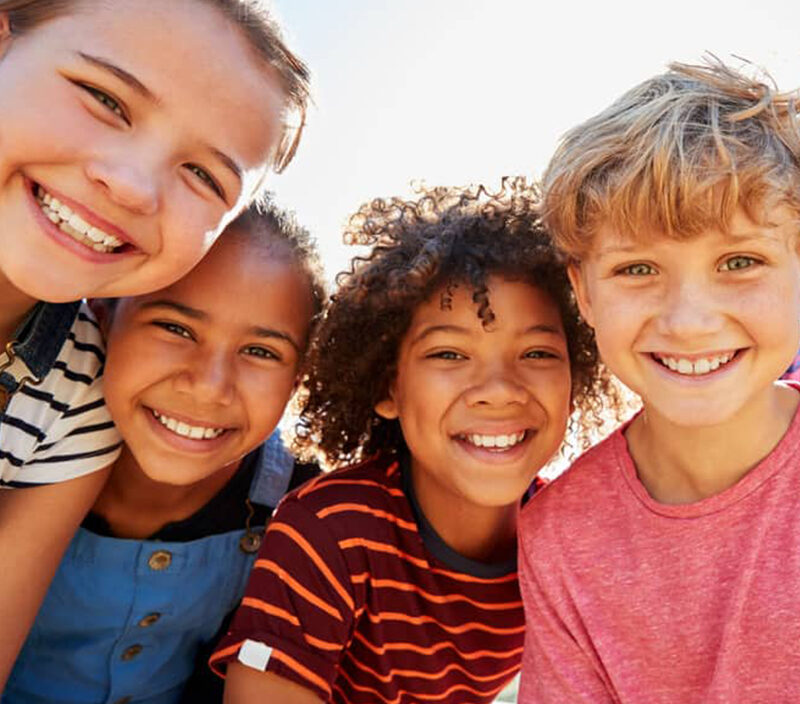 Four children smiling at the camera outdoors on a sunny day.