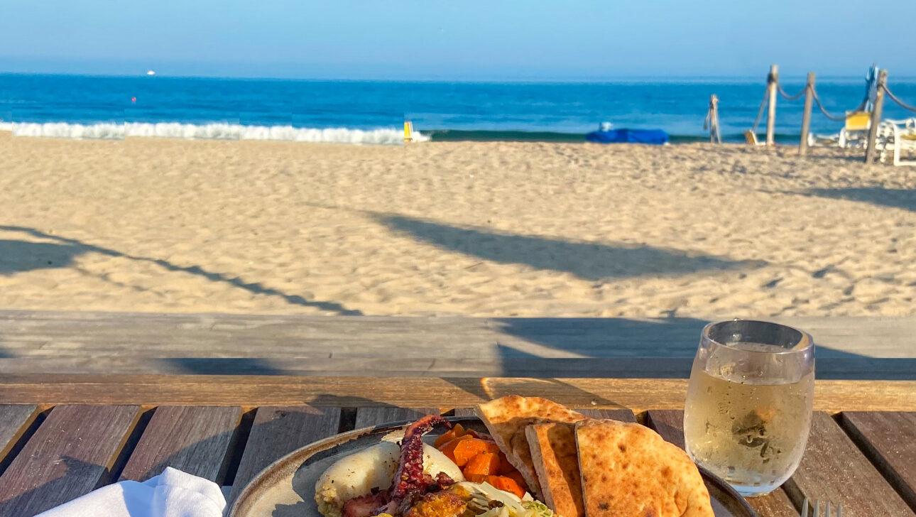 A plate of assorted grilled vegetables and flatbread on a wooden table by the beach, with a glass of water and a napkin.