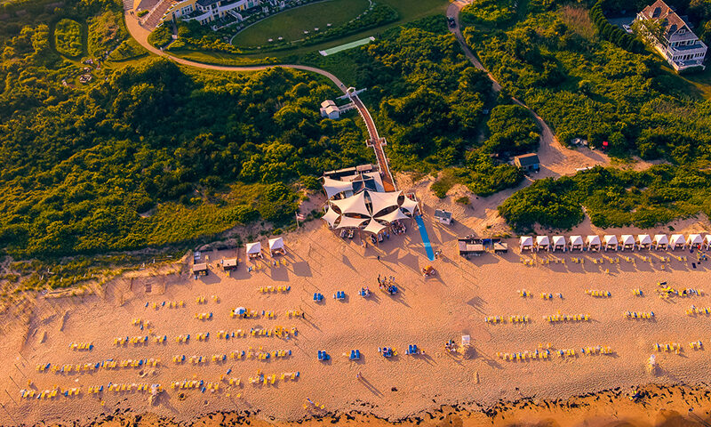 Aerial view of a beachfront resort with rows of loungers and umbrellas on the sand, surrounded by green landscape and several buildings in the background. Waves approach the shore.