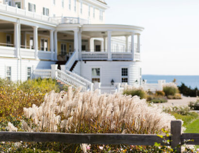 A grand white building with multiple balconies overlooks a garden with ornamental grasses, with the ocean visible in the background.