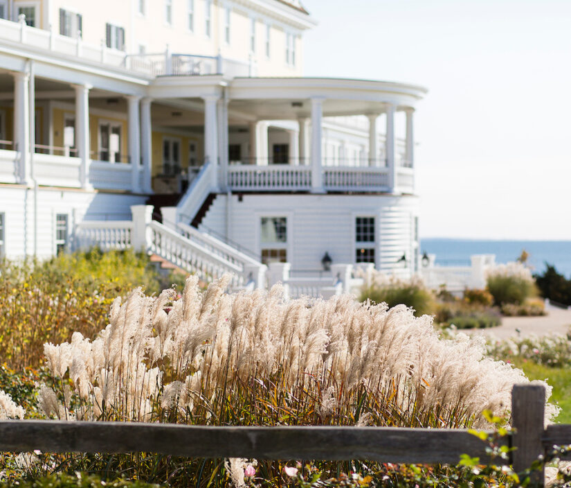 A grand white building with multiple balconies overlooks a garden with ornamental grasses, with the ocean visible in the background.