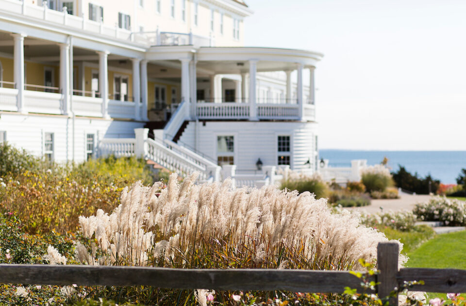 A grand white building with multiple balconies overlooks a garden with ornamental grasses, with the ocean visible in the background.