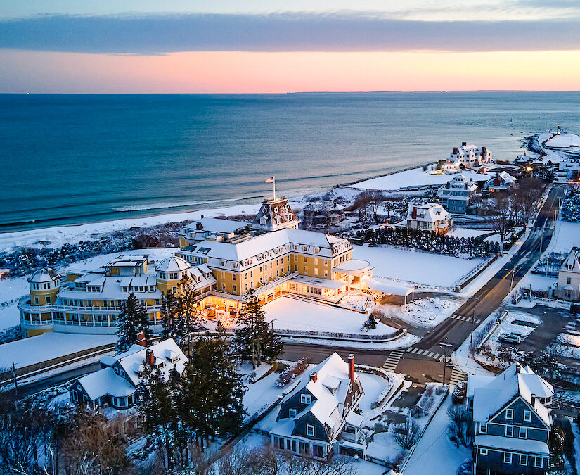 Aerial view of a coastal town with snow-covered buildings, roads, and a beach at sunset. A large building with lit windows is prominent in the foreground.