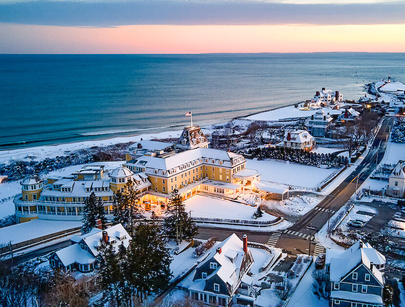 Aerial view of a coastal town with snow-covered buildings, roads, and a beach at sunset. A large building with lit windows is prominent in the foreground.