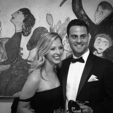 A couple in formal attire poses in front of a wall with abstract art in the background.