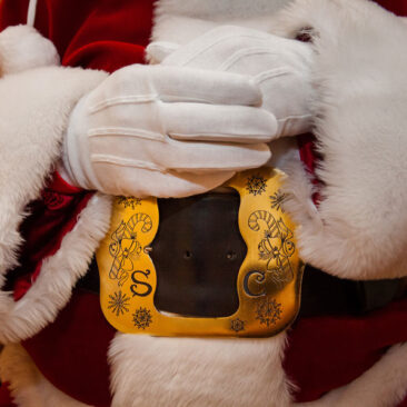 A person dressed in a red and white fur-trimmed costume with white gloves, featuring a large gold belt buckle with decorative designs.